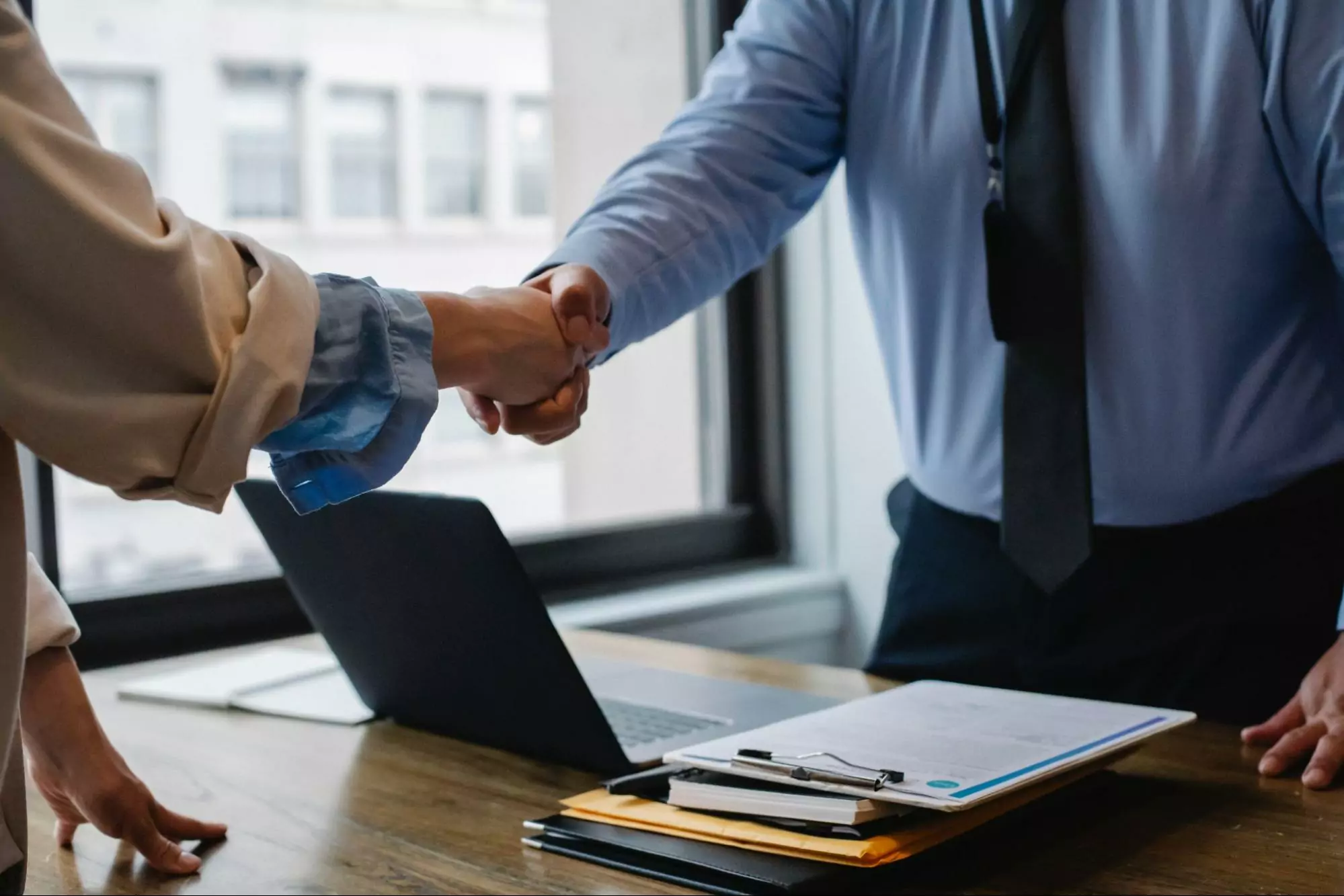 Two professionals shaking hands over a desk with documents and laptop.